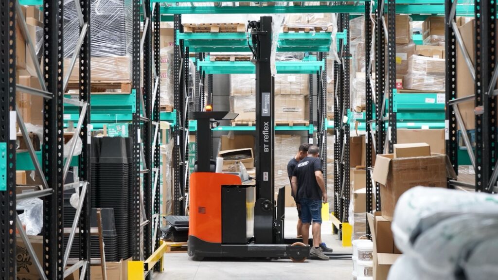 3PL warehouse workers beside a forklift in an aisle surrounded by storage racks, boxes, and palletized goods.